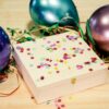 Weiße Holzbox mit bunten Konfetti-Elementen, umgeben von Ballons und farbigen Bändern auf Holzuntergrund