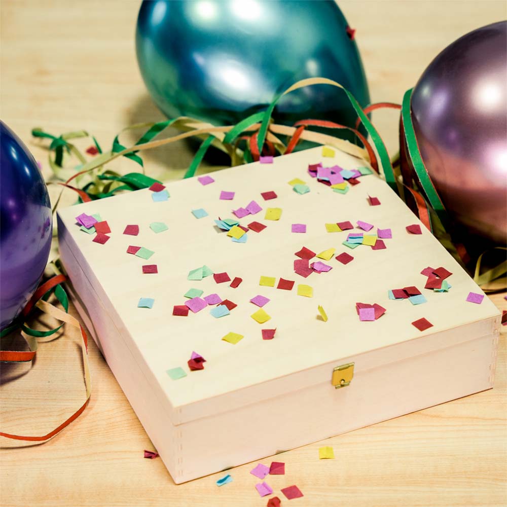 Weiße Holzbox mit bunten Konfetti-Elementen, umgeben von Ballons und farbigen Bändern auf Holzuntergrund
